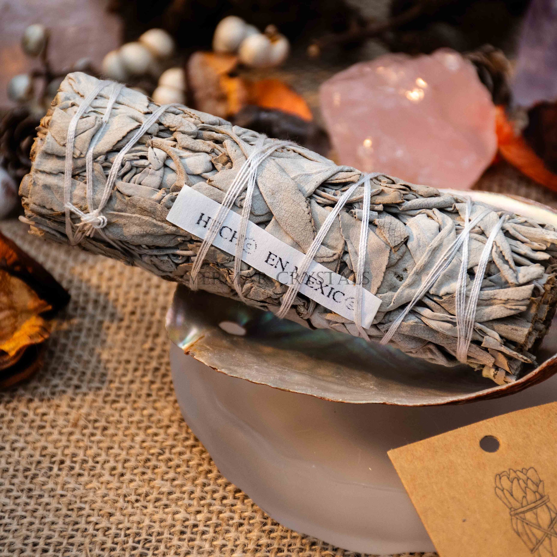 Close-up of a tightly bundled white sage smudge stick with label resting on a shell and rustic fabric background