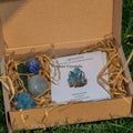 Open cardboard box containing blue and green polished crystals with a card labeled Apatite, stone of manifestation, on shredded paper filling