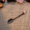 Handcrafted antique bronze tone alchemy spoon with floral engravings and rose quartz crystal on burlap background