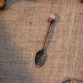 Handcrafted bronze-tone alchemy spoon with floral engravings and pink crystal tip on burlap background