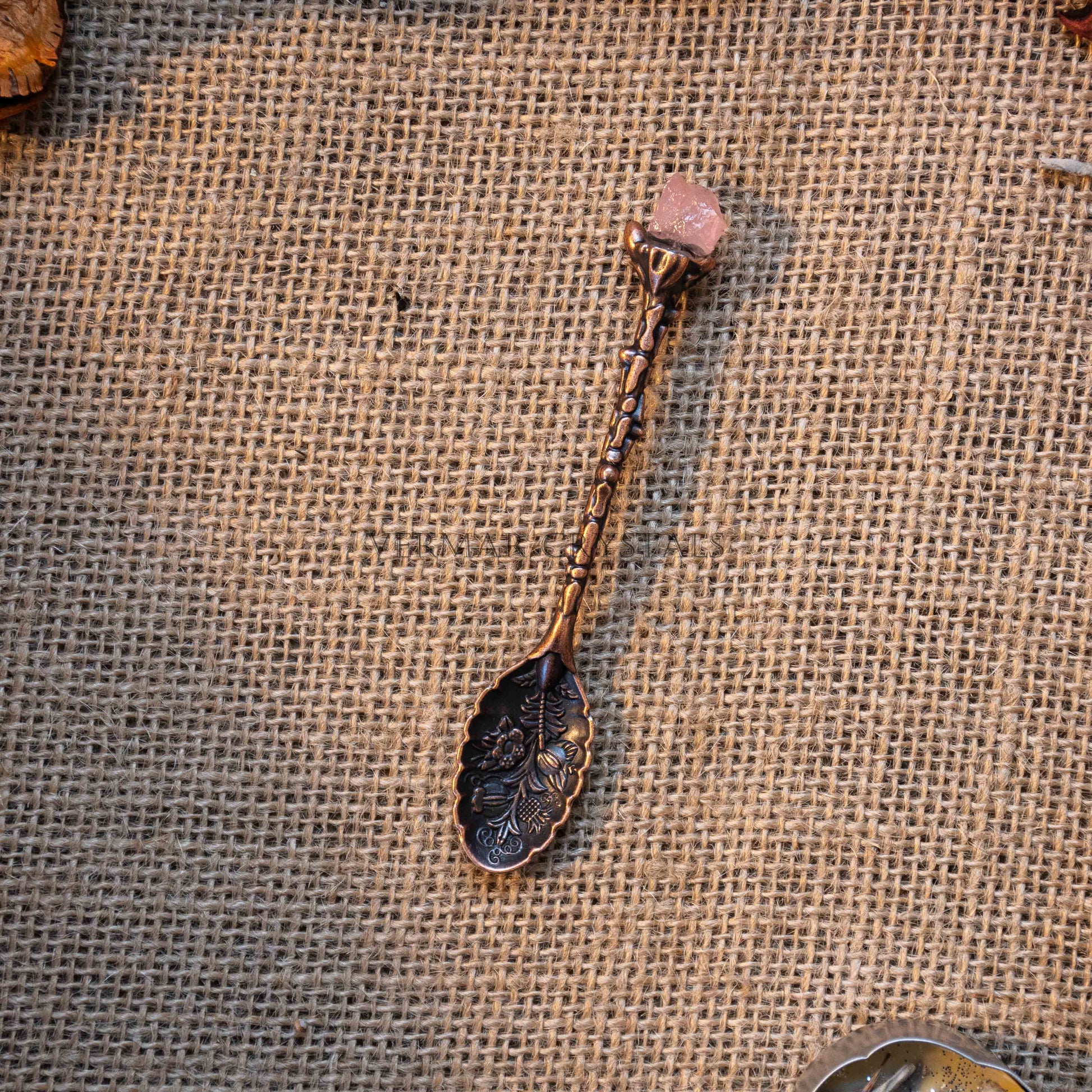 Handcrafted bronze-tone alchemy spoon with floral engravings and pink crystal tip on burlap background