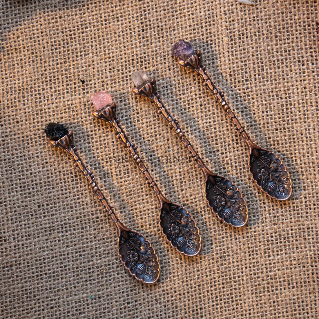 Four antique bronze tone alchemy spoons with floral engravings and polished crystal handles on burlap background