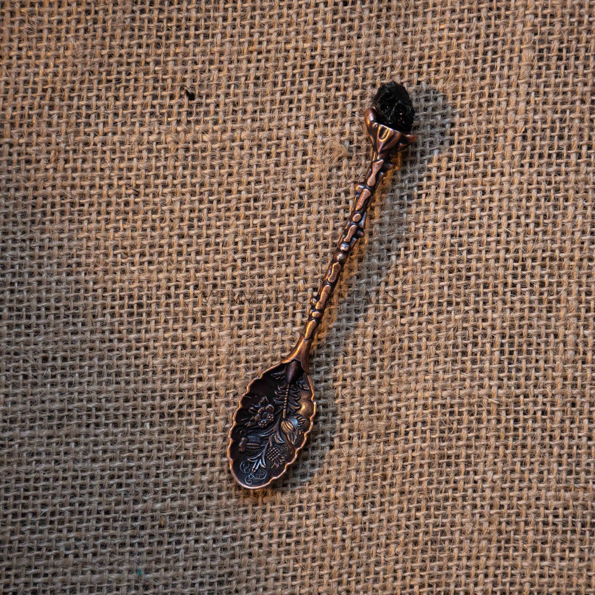 Antique bronze tone handcrafted spoon with floral engravings and black crystal handle on burlap fabric background