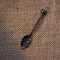 Antique bronze-toned alchemy spoon with floral engravings and a crystal tip resting on burlap fabric background