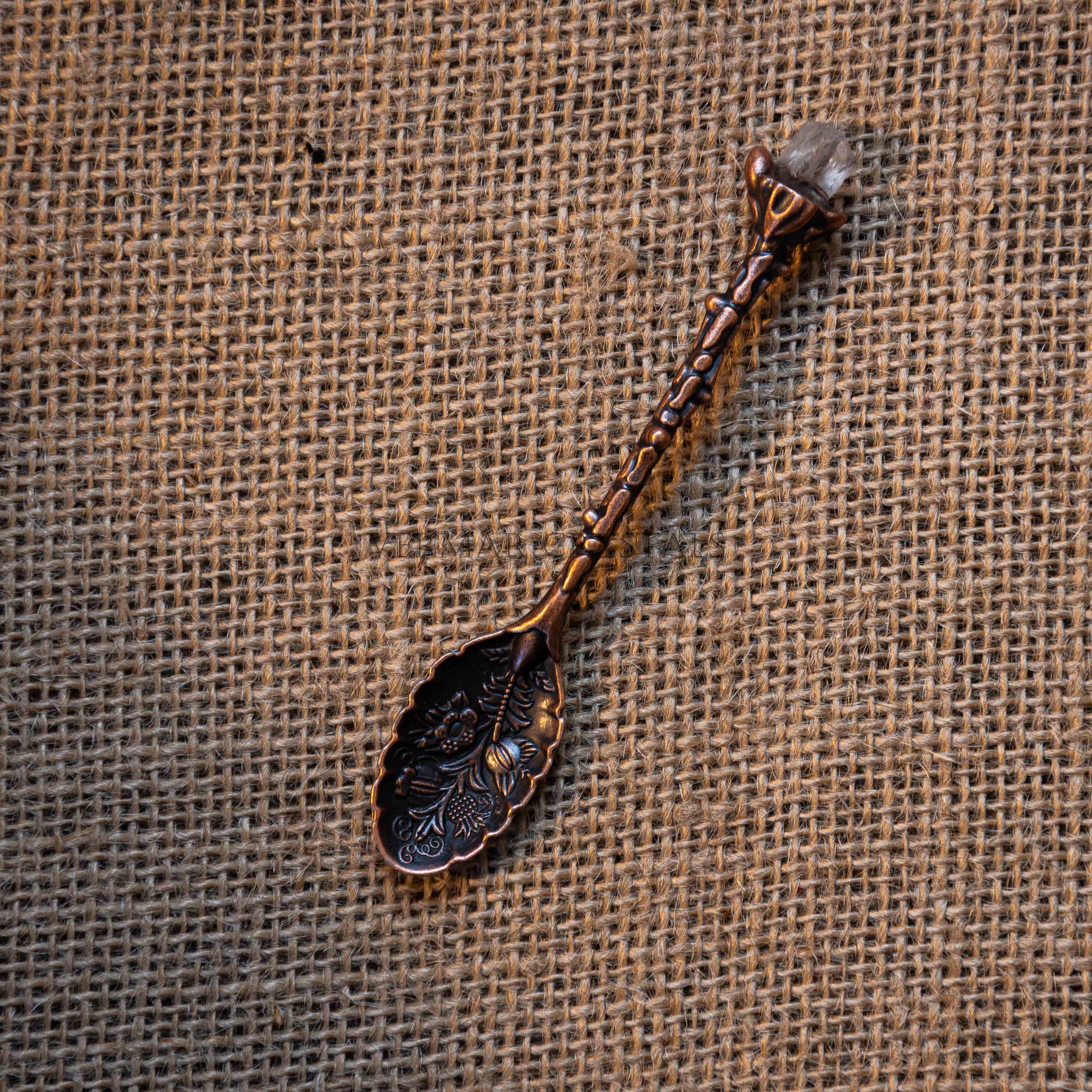 Antique bronze-toned alchemy spoon with floral engravings and a crystal tip resting on burlap fabric background