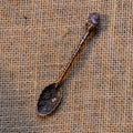 Antique bronze-toned alchemy spoon with floral engravings and a small amethyst crystal on burlap fabric background