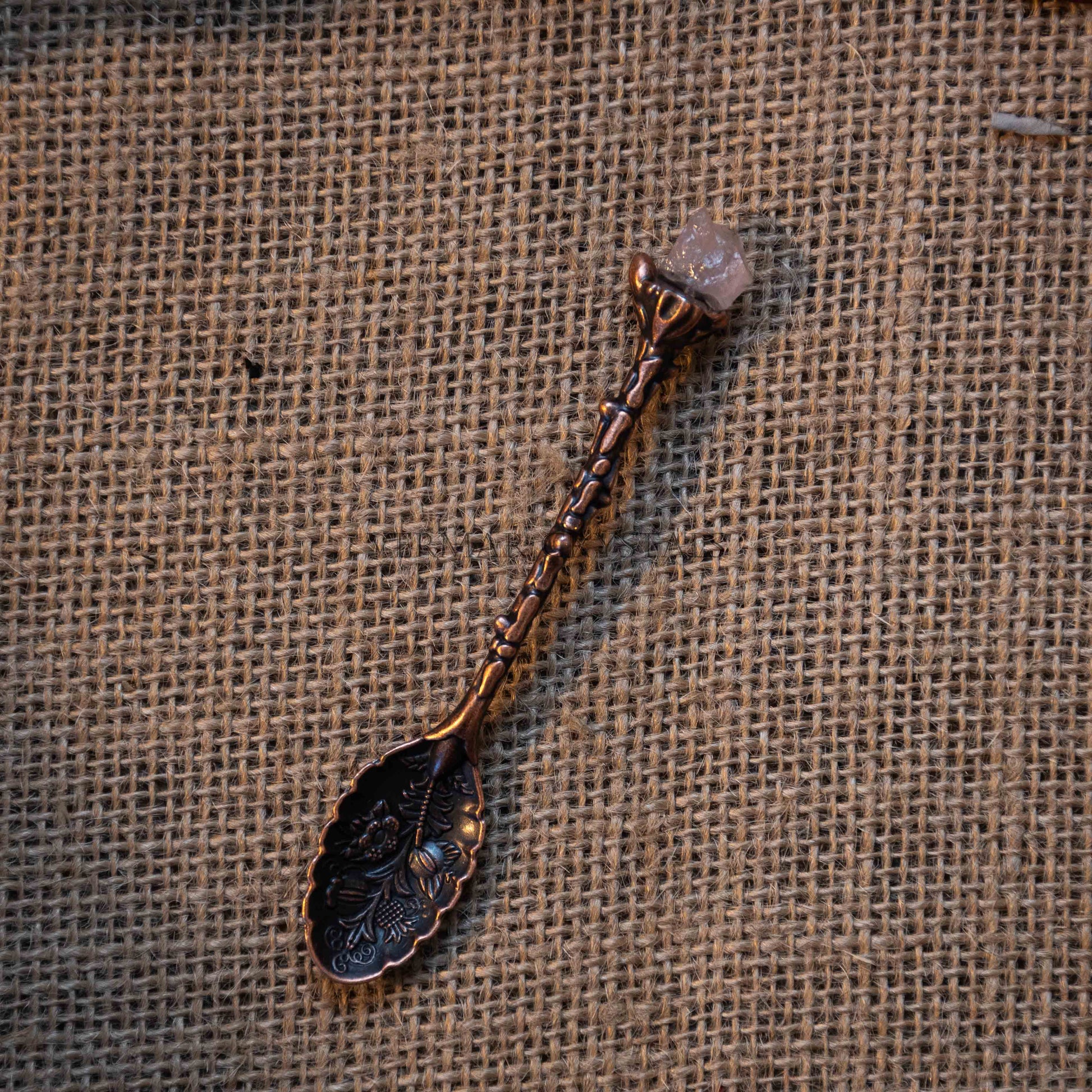 Antique bronze alchemy spoon with floral engravings and raw rose quartz crystal on rustic burlap background