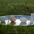 Set of polished and raw crystals including light green, peach, and white stones on a silver tray outdoors on grass background
