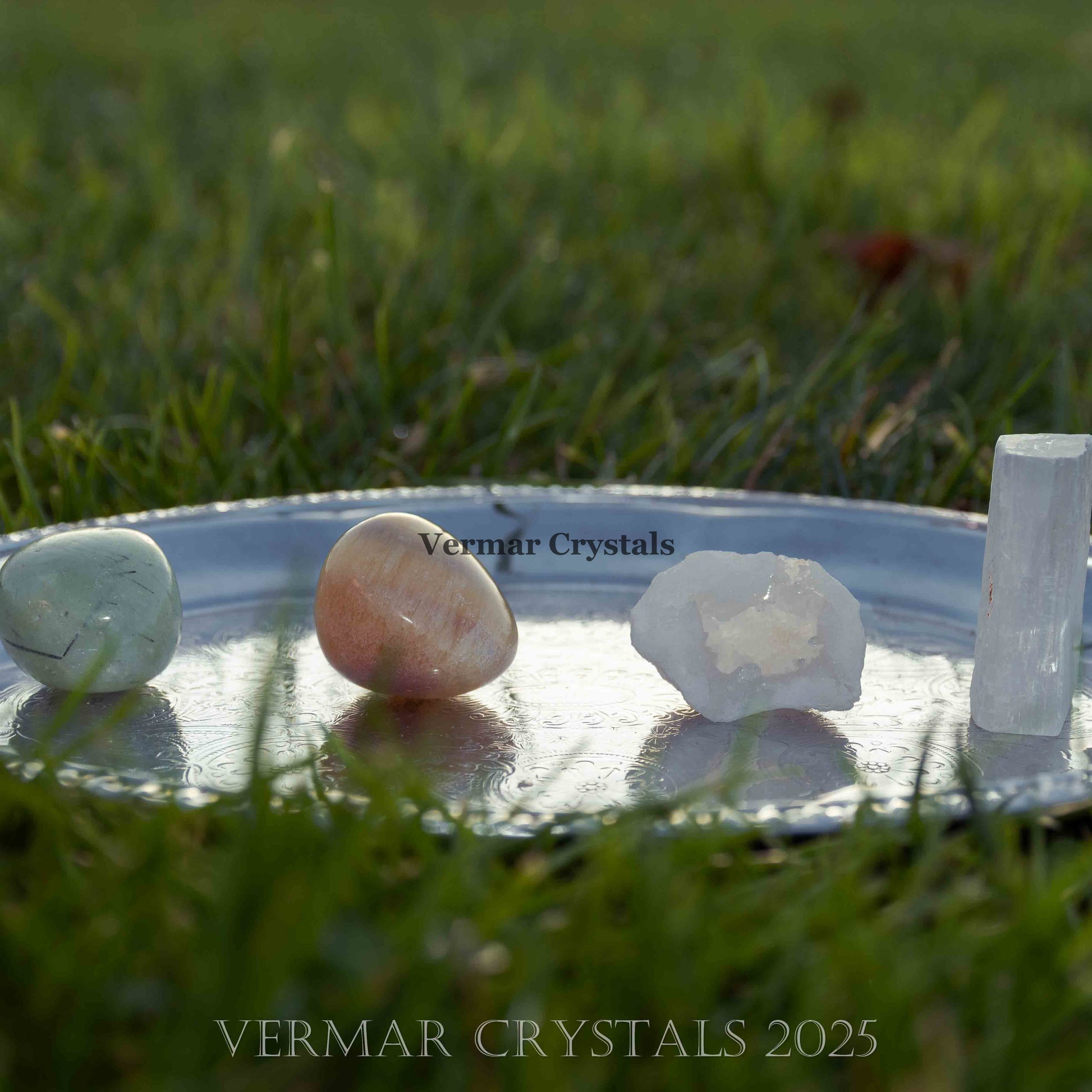 Set of polished and raw crystals including light green, peach, and white stones on a silver tray outdoors on grass background