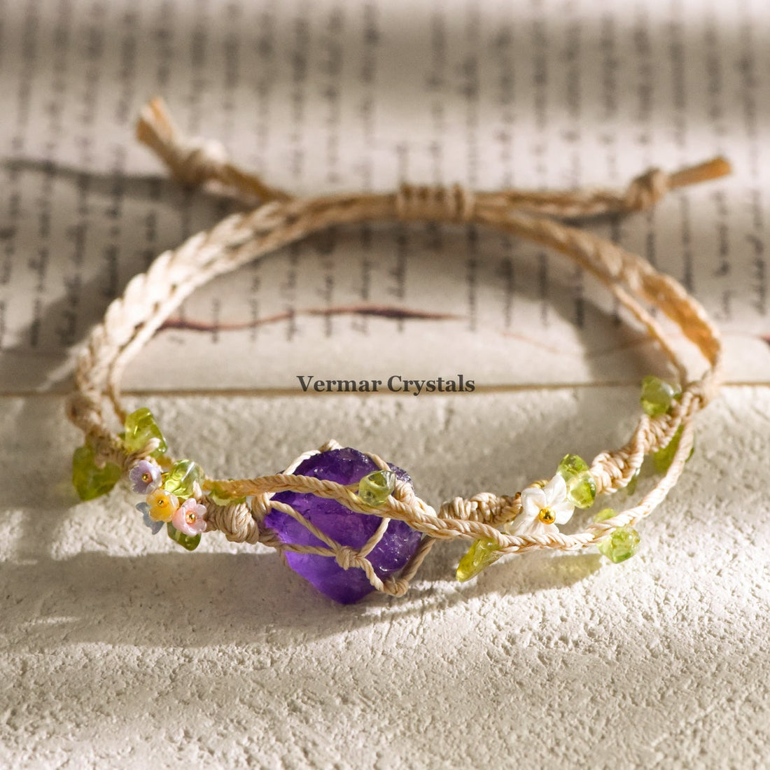 Handcrafted amethyst stone wrapped in braided cord with small flower and green bead accents on textured surface