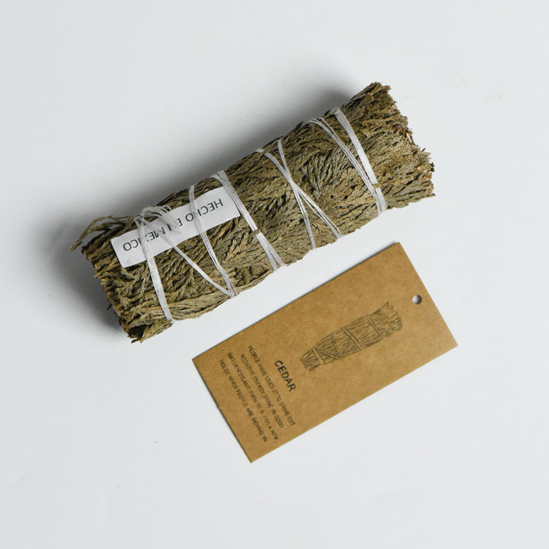 Natural cedar smudge stick tied with white string alongside a brown tag with cedar illustration on white background