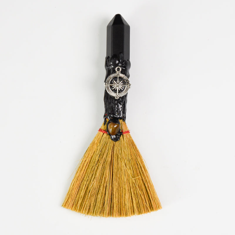 Handcrafted ritual witch broom with natural straw, black crystal handle, compass charm, and tiger's eye stone on white background