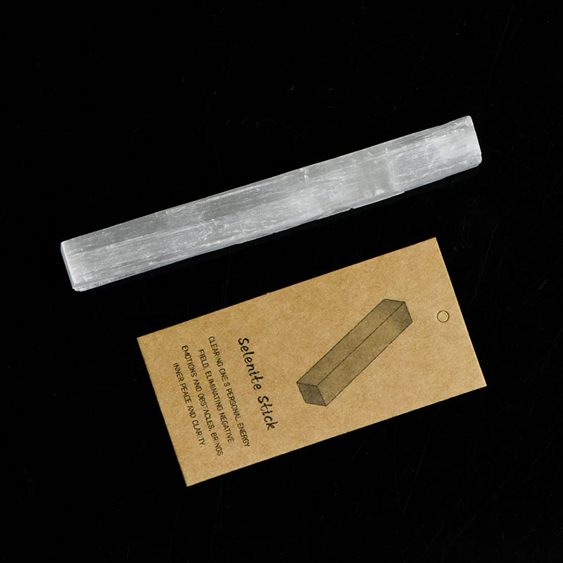 Natural 10cm selenite stick crystal next to a brown kraft paper tag on solid black background