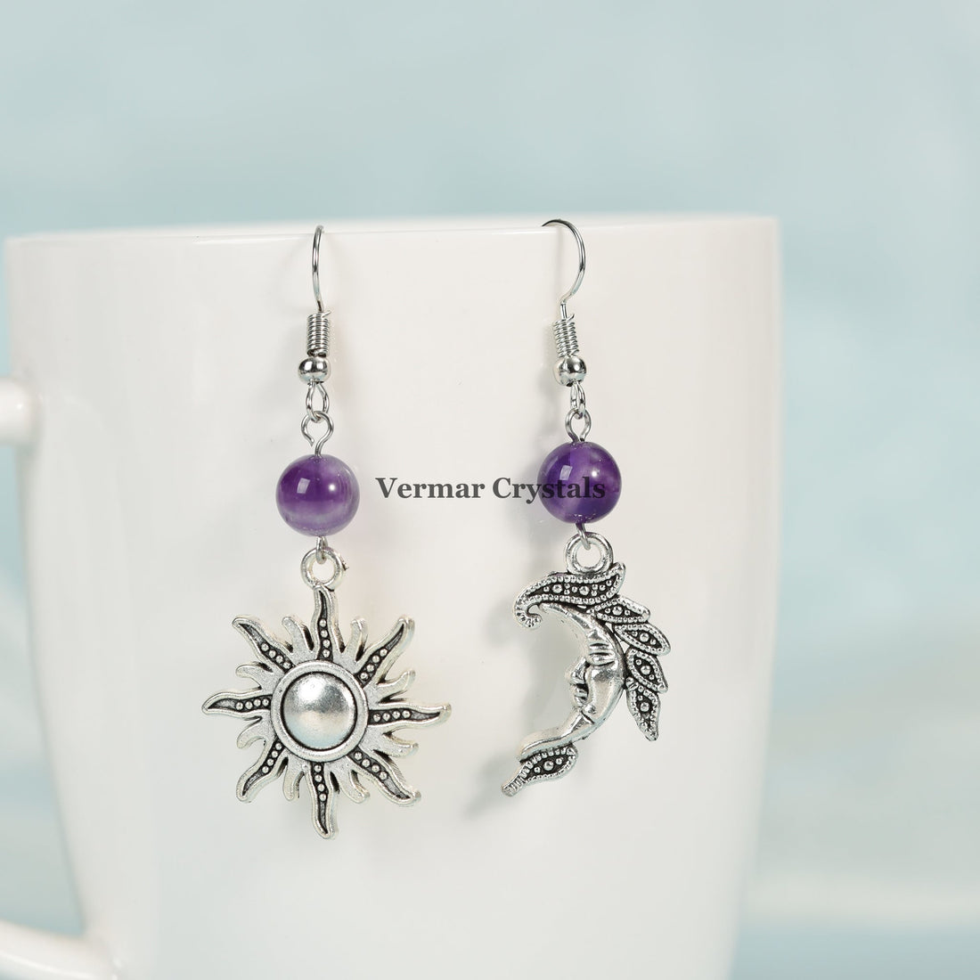 Asymmetric sun and moon silver earrings with polished amethyst beads hanging against white cup background
