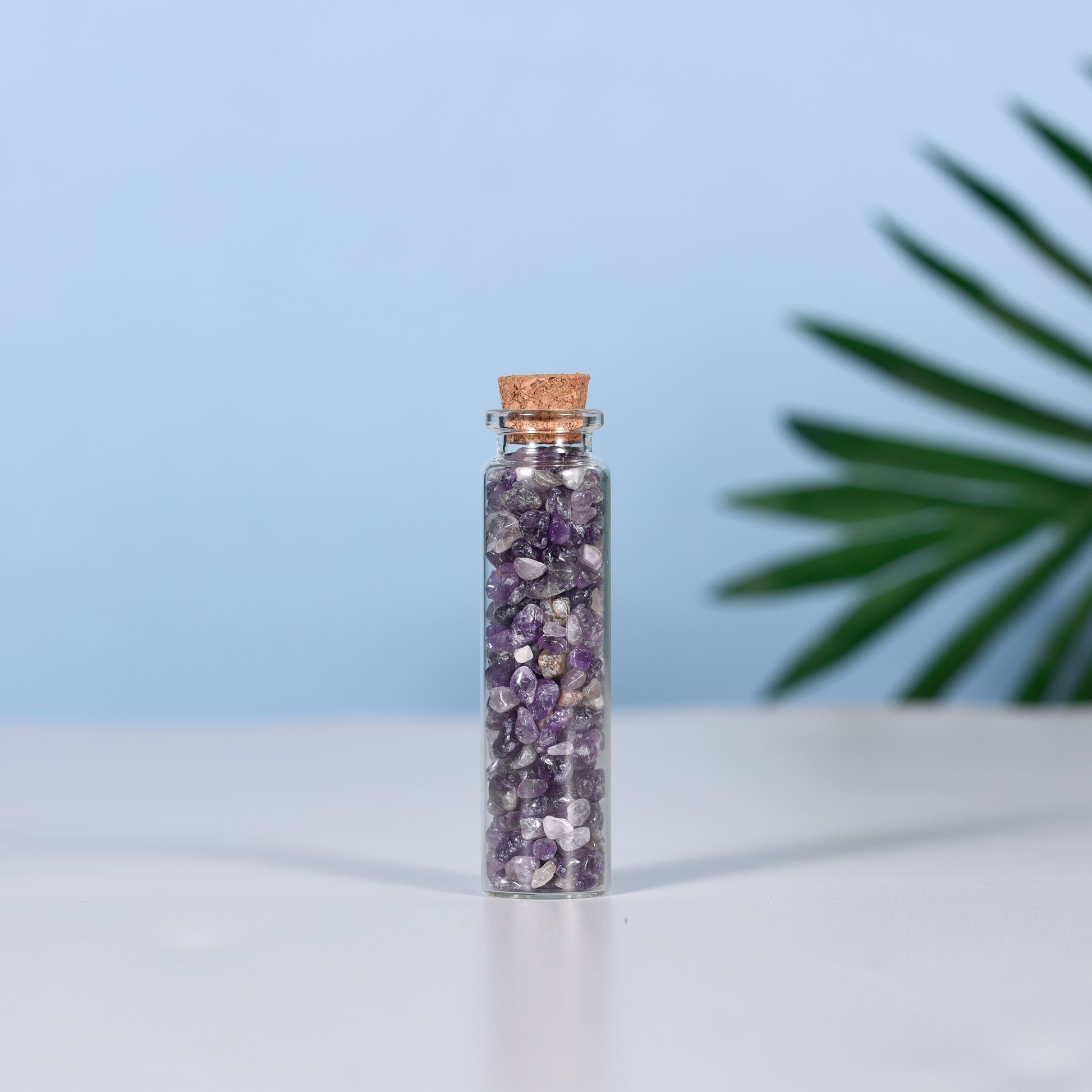 Mini glass vial filled with natural purple amethyst crystal chips sealed with cork on white surface and blue background