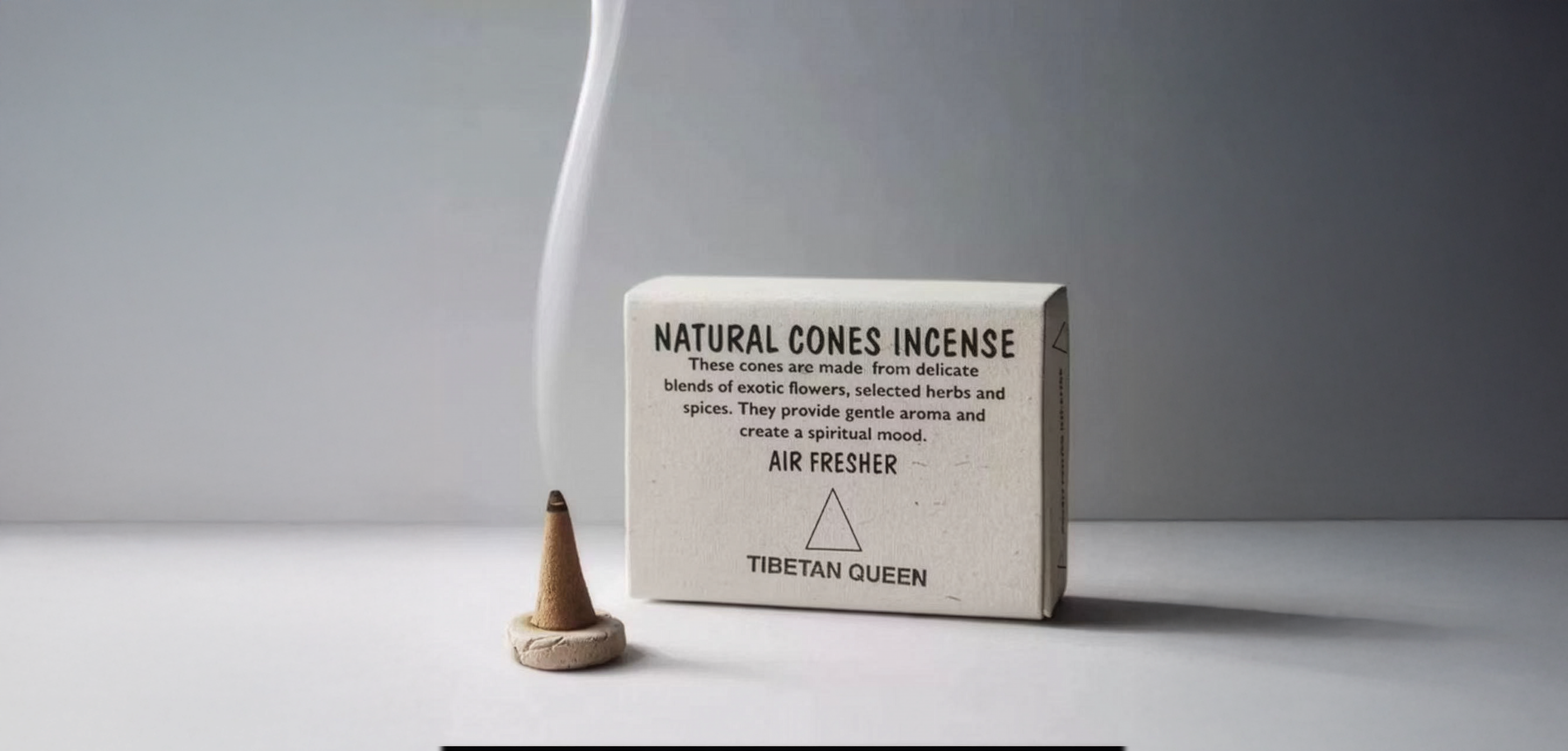 Single lit natural incense cone releasing smoke next to a rectangular box labeled Tibetan Queen on white surfaceslider_item_f44eqh