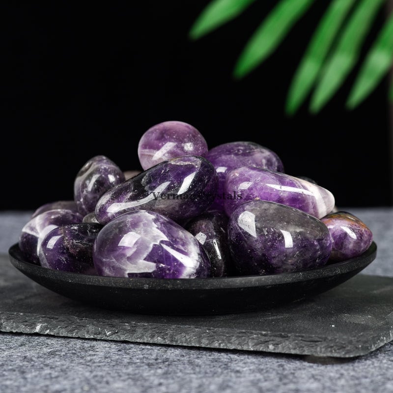 Black dish filled with smooth polished amethyst tumblestones in various shades of purple on a dark surface