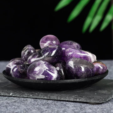 Black dish filled with smooth polished amethyst tumblestones in various shades of purple on a dark surface