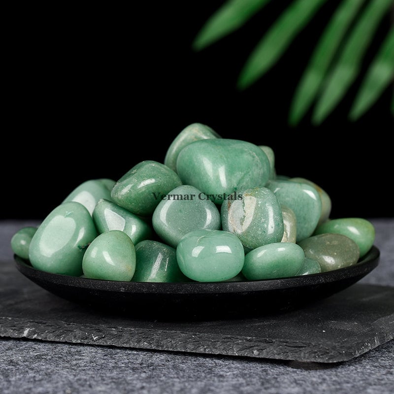 Polished green aventurine tumblestones arranged on a black dish with a dark background and green leaves above