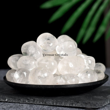 Pile of polished clear quartz tumblestones displayed on a black dish with a dark background and green leaf detail